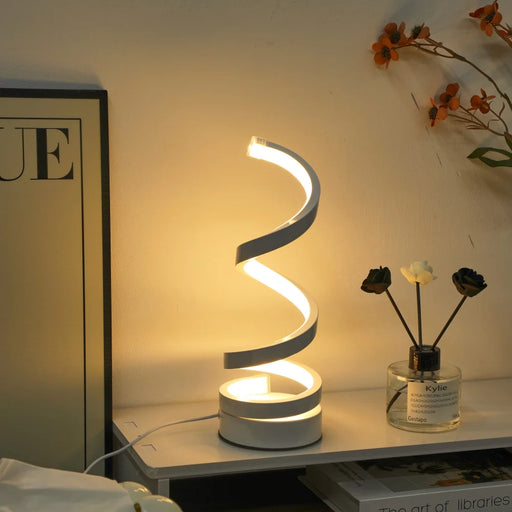 Modern spiral LED lamp on a bedside table with a magazine and small plant.